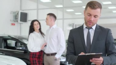 Portrait of a male car sales manager. In the background, customers choose a car.