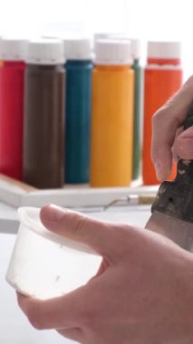 Kapatın. Ressamların eli, kaptaki beyaz boyayı spatulayla karıştırır. Beyaz tonlarda yaratıcı atölye çalışması. Bir resim yaratma süreci. Fırça hareketi. Dikey video