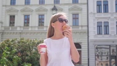 Şehir yürüyüşünde kruvasan ve bir fincan kahveyle genç bir kadın. Güneş gözlüklü güzel sarışın bir kadın bir fincan kahve tutuyor ve kruvasan yiyor..