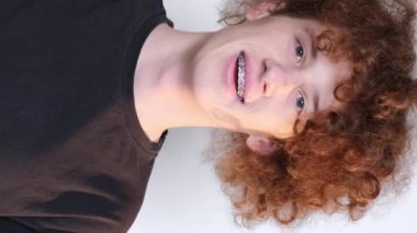 Face of satisfied crazy happy young guy with braces showing thumbs up. Positive curly red-haired student on a light blue background. Vertical video