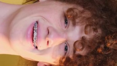 Close-up of the face of a satisfied, smiling young guy with braces. Positive curly red-haired student on a brown background. Vertical video