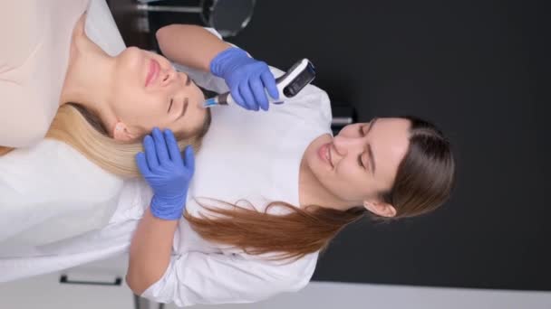 垂直录像 美容师为病人进行面部电按摩 一个女人在美容院里一个年轻的女人接受电按摩 — 图库视频影像