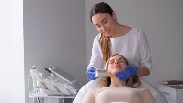 化粧品クリニックで顔の治療プロセス中に患者とプロの美容師 若い女性が電動フェイシャルマッサージを受ける しわの肌の若返りと滑らかさ — ストック動画