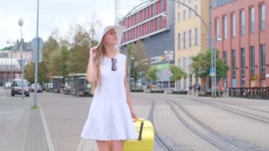 Beyaz şapkalı, bavullu bir kadın tramvay raylarının yakınından geçiyor..