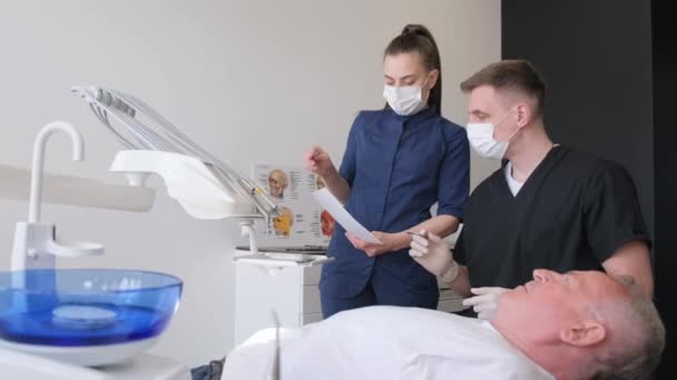 一名男牙医向他的老年病人解释植入物是如何固定的 一位老人坐在牙椅上听着 牙医的工作 — 图库视频影像