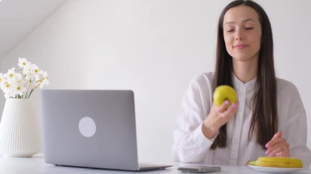 一个黑发女人在家里的电脑前工作 吃着一个苹果 工作时的健康小吃 — 图库视频影像