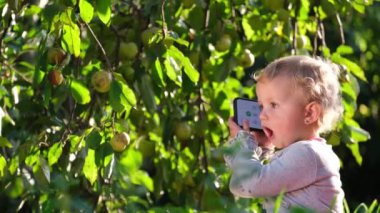 Şirin, mutlu, gülümseyen iki yaşında bir kız bahçedeki yeşil çimenlerde oturuyor, akıllı telefonuyla oynuyor ve gülüyor..