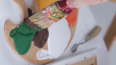 The hand of a young artist mixes paints on a palette that stands on the table. Basic colors. Work with a thick paintbrush. Workplace of the artist