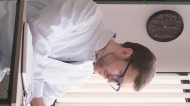 Gözlüklü ciddi bir doktor, dizüstü bilgisayarda çalışıyor, ofiste bir masada oturuyor, beyaz üniformalı bir terapist. Dikey video