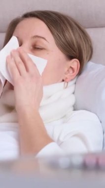 Soğuk algınlığı olan Avrupalı bir kadın bandajlı bir boğazla yatakta yatıyor. Evde mevsimsel grip ve soğuk algınlığı tedavisi. Dikey video