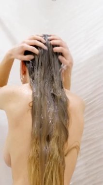 Sıcak bir duş deryasının altında genç bir kadın elleriyle saçlarına masaj yapıyor. Kafasını şampuanla yıkamak. Güzel, uzun saçlara dikkat et. Dikey video