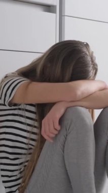 Mutfak odasında yerde oturan yorgun genç bir kadın, başı ağrıyor ve üzgün, üzgün, mutsuz hissediyor. Dikey video