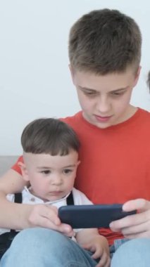 Farklı yaşlarda üç çocuk kanepede otururken akıllı telefon kullanıyor. Çocuklar çerçevenin merkezini işgal ediyor. Işık sıcak ve tekdüze. Dikey video