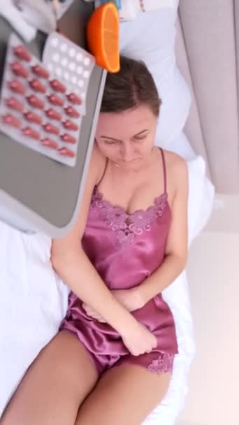 一个穿着漂亮睡衣的年轻女人躺在床上 痛苦不堪 月经期疼痛 用药片治疗疼痛 垂直录像 — 图库视频影像