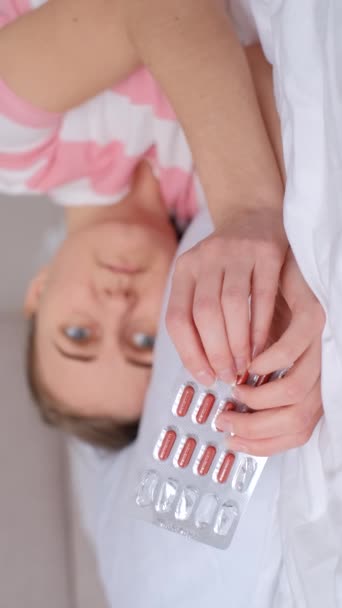胃の痛みを伴う女性は鎮痛剤を飲んでベッドに横たわっています 痛ましい期間 女性の月経周期 バーティカルビデオ — ストック動画