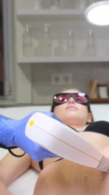 Kız güzellik uzmanının ofisinde yatıyor, doktorlar ellerini bacağına bir lazer cihazıyla sürdüler. Üniforma ve eldivenler. Dikey video