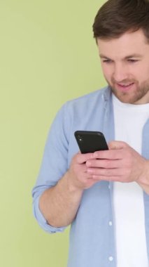 Stüdyo arka planında telefonu olan bir adam kot gömlek giyiyor ve cihazının ekranına bakmakla meşgul ve mesajlarıyla çok ilgileniyor. Dikey video