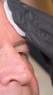 Kozmetik uzmanı tarafından muayene edilen yaşlı bir adam modern bir klinikte bir sandalyede oturuyor. Kırışıklıkların tedavisini enjeksiyonla yapmak. Dikey video