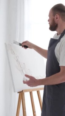 Profesyonel bir sanatçı beyaz bir tuvale metal bir fırça ve sehpa kullanarak resim çizer. Bir sanatçının alçı tablosu yaratma süreci. Dikey video