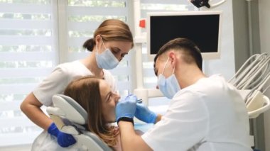 Bir dişçi ve bir asistan kadın bir hastanın dişlerini muayene eder. Modern bir klinikte diş tedavisi. Dikey video