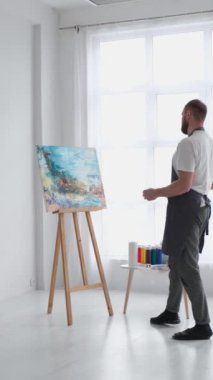 Yetenekli bir erkek sanatçı, fırça kullanarak, beyaz bir tuvalde yağlı boya tablosunun modern bir başyapıtını yaratıyor. Tuval büyük bir stüdyoda bir sehpanın üzerinde. Dikey video