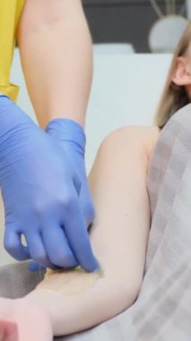 Sarı takım elbiseli bir güzellik uzmanı genç bir müşterinin elini tutuyor. Tüy dökümü için şeker ezmesi. Acısız bir epilasyon. Güzel bir vücudu önemsemek. Dikey video