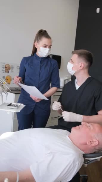歯科医は 歯医者を訪問する上級男性のアシスタントと協力し 診療所 歯科コンセプトで歯を調べています バーティカルビデオ — ストック動画
