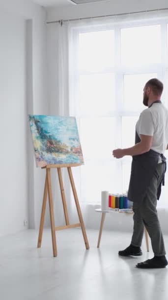 才能ある男性アーティストは ブラシを使用して 白いキャンバスに油絵の現代的な傑作を作成します キャンバスは大きなスタジオのイーゼルにある バーティカルビデオ — ストック動画
