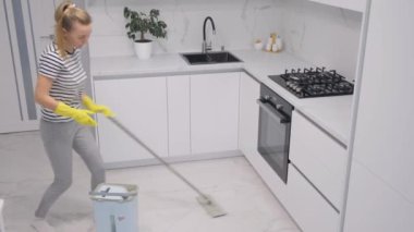 Güzel, çekici, neşeli kız seramik yerleri paspaslıyor, dans ediyor, modern beyaz ışık iç mekan mutfağında eğleniyor..