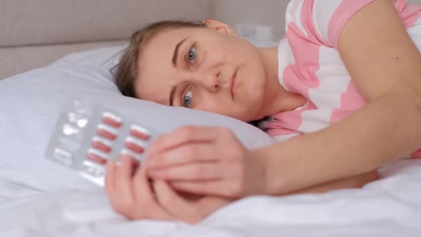 病気の女性がベッドに横になり 薬を手に持っている 家庭でのウイルス性および細菌性疾患の治療 — ストック動画