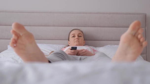 ある女性がテレビのリモコンを持っていて 寝室の広いベッドに横たわっている 疲れた女性 バーティカルビデオ — ストック動画