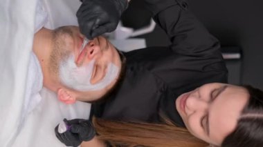 Kaplıcada yüz bakımı yaptıran bir adamın dikey videosu. Bir kozmetolog yüzüne kilden bir maske takarak yüz masajı yapar. Dikey video