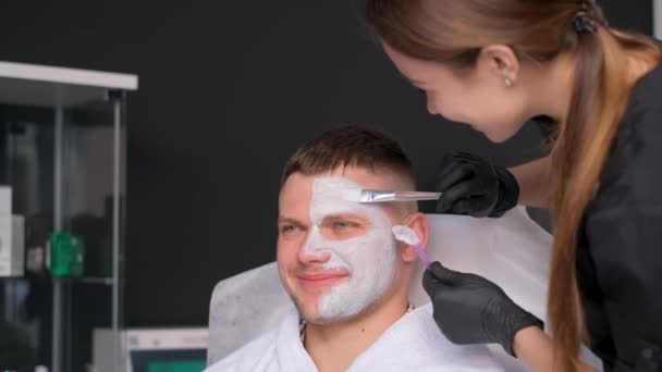スパの若い男性が顔の治療を受けています 人の顔に粘土のマスク 化粧品師は顔に保湿マスクを施したフェイシャルマッサージを行います バーティカルビデオ — ストック動画