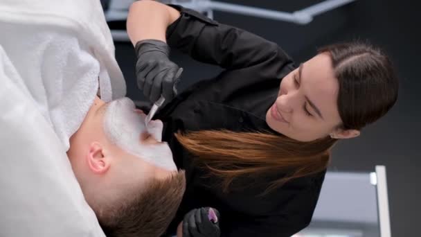 美容师在由一位女性美容师进行的面部清洁过程中的英俊男子恢复粘土面罩 垂直录像 — 图库视频影像