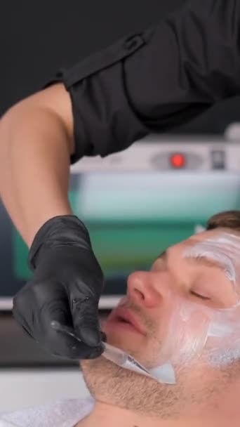 男性面部整容和抗衰老美容术 美容师在由一位女性美容师进行的面部清洁过程中的英俊男子粘土使面膜年轻化 垂直录像 — 图库视频影像