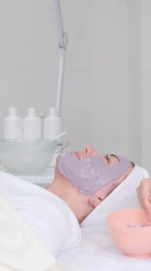 Bir kadın kilden bir maskeyle rahatlar, bir güzellik uzmanı müşterinin yüzüne bir fırça sürer. Modern kozmetoloji spa salonu. Dikey video