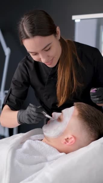 美容师在由一位女性美容师进行的面部清洁过程中的英俊男子恢复粘土面罩 垂直录像 — 图库视频影像