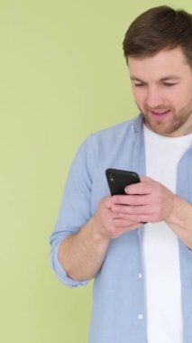 Cep telefonunu kullanan genç adam yeşil arka planda mutlu ve mutlu görünüyor. Modern teknolojiyi elinde tutan, yumruklarını kaldıran, evet diye bağıran neşeli bir adam. Dikey video