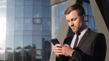 Siyah takım elbiseli gülümseyen genç bir işadamı akıllı telefona bakıyor. İş merkezinin yakınında elinde cep telefonu olan bir yönetici. İş konsepti