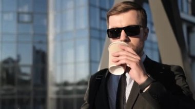 Sakallı ve siyah gözlüklü genç bir iş adamı güneşli bir günde dışarıda kahve içiyor. Arka plandaki cam gökdelen yöneticisi