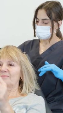 Tatmin olmuş yaşlı bir kadının trichologla olan randevusunun dikey videosu. Genç bir doktor hastaların kafa derilerini tarıyor. Dikey video