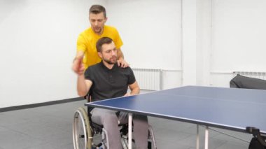 Rehabilitasyon doktoru tekerlekli sandalyedeki birinin masa tenisi oynamasına yardım eder.. 