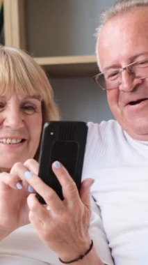 Büyükbabam ve büyükannem evde kanepede oturmuş akıllı telefondan eğlence programları izliyorlar. Gülümseyen emekli aile çifti. Dikey video