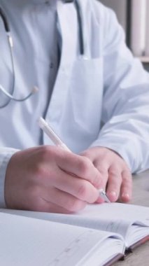 Gözlüklü ve üniformalı profesyonel genç doktor ofisinde evrak işleriyle uğraşıyor. Teşhis, tedavi planı yazıyorum. İş yerinde bir doktor. Dikey video
