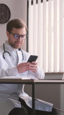 Ofisteki masada oturan genç profesyonel doktor akıllı telefona bakıyor. Doktorlar gündüzleri çalışıyor. Özel terapist muayenehanesi. Dikey video