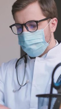 Tıbbi maskeli ve gözlüklü genç bir doktor ofisinde bir netbook üzerinde çalışıyor. Fonendoskoplu bir Infersionist doktor. İşteki doktor. Dikey video