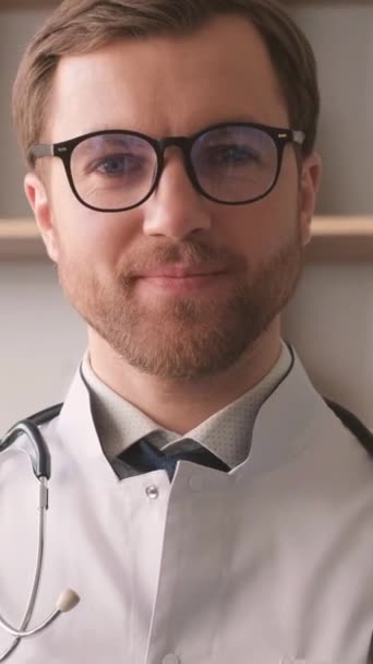 白い制服と本棚の背景に立っているハンサムな若い医師の肖像画 セラピスト医師の肖像画 バーティカルビデオ — ストック動画