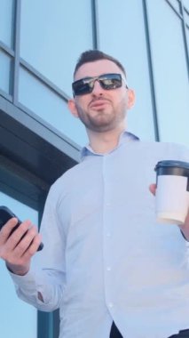 Yakışıklı bir adam gökdelenin yanında duruyor, kahve içiyor ve kamerayla konuşuyor. Başarılı bir stil sahibi iş adamı sokakta onunla kahve içiyor. Dikey video