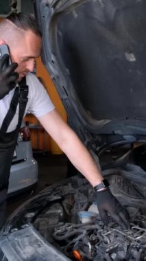 Avrupalı tamirci telefonda konuşuyor. Araçların bakımı. Bir atölyede araba motoru tamiri. Dikey video