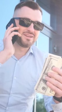 Elinde dolarlar olan genç bir patron akıllı bir telefonla konuşuyor. İşletme müdürü konsepti. Dikey video